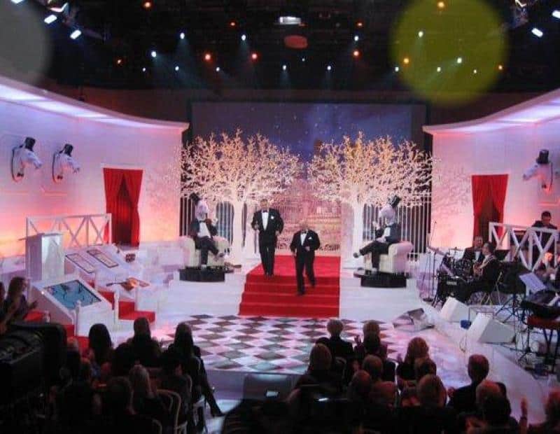 oryg_064490685_1129949550544944_1465385417682452480_o Scena z występu na żywo z dekoracjami w stylu glamour, dwaj mężczyźni w garniturach na czerwonym dywanie, widownia i zespół muzyczny.