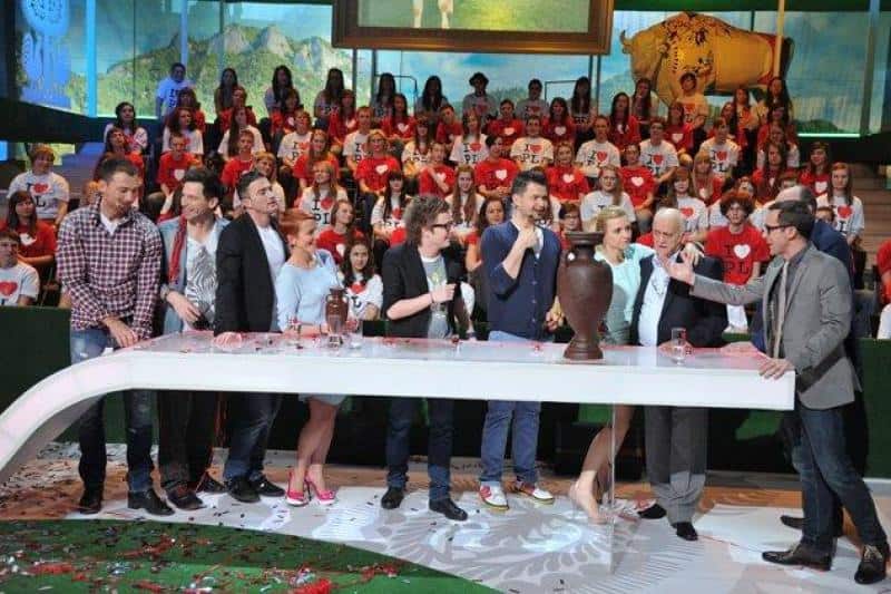 oryg_064490685_1129949550544944_1465385417682452480_o Grupa osób stojąca przy stole w studiu telewizyjnym; w tle widownia ubrana w białe i czerwone koszulki.