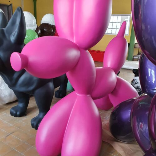 Jasnoróżowa rzeźba balonowego psa w pomieszczeniu, w tle inne rzeźby.