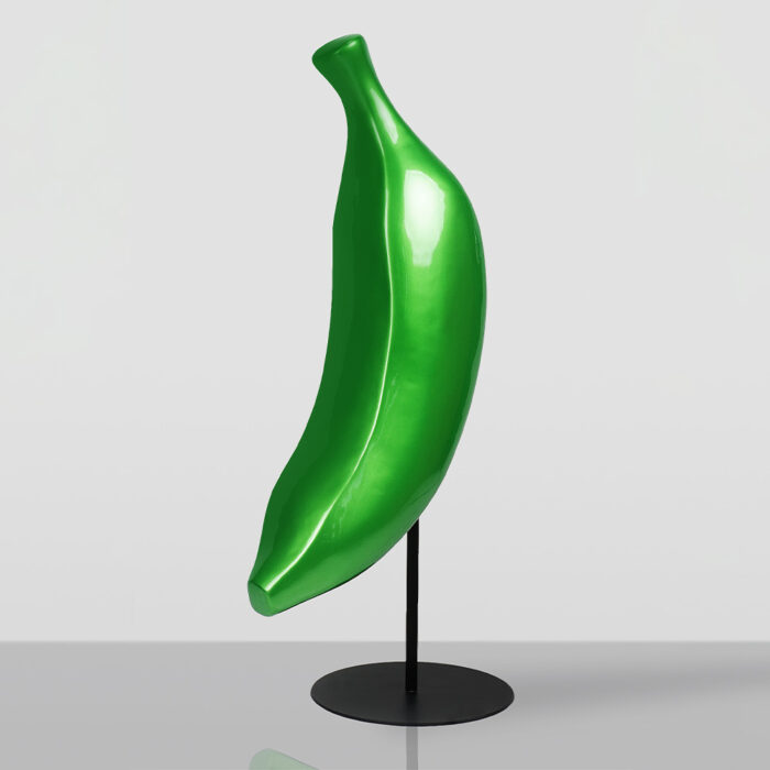 Rzeźba zielonego banana na czarnej podstawie stojącej na szarej powierzchni na tle jasnej ściany.