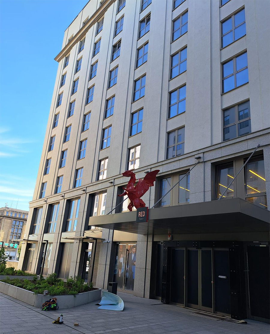 Smoki Low Poly w Hotelu Radisson 8 Wysoki budynek z szarymi ścianami i licznymi oknami, przed wejściem czerwony posąg smoka z napisem "RED".