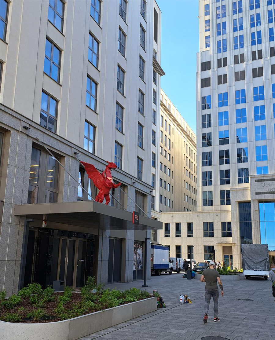 Smoki Low Poly w Hotelu Radisson 5 Budynek z czerwoną rzeźbą konia na daszku oraz osoba przechodząca chodnikiem.