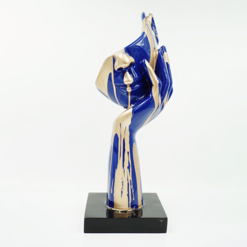 Rzeźba przedstawiająca dłoń z twarzą w odcieniach niebieskiego i złotego, stojąca na czarnej podstawie na białym tle