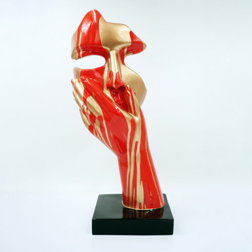 Rzeźba przedstawiająca czerwoną i złotą abstrakcyjną formę dłoni podtrzymującej fragment twarzy, stojąca na czarnej podstawie na białym tle.