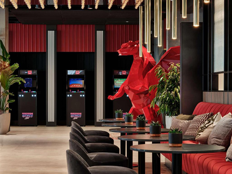 Smoki Low Poly w Hotelu Radisson 3 Wnętrze nowoczesnego salonu z czerwoną ławą, czarnymi fotelami, stylowymi stołami i dużą czerwoną rzeźbą smoka, w tle automaty do gier retro.