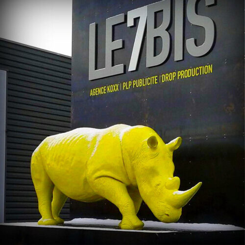 żółta rzeźba nosorożca stojąca przed ścianą z napisem LE7BIS oraz mniejszymi napisami agencji reklamowej