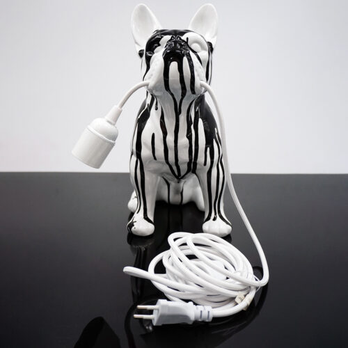 Ceramiczna figurka psa w czarno-białe paski trzymająca w pysku biały kabel elektryczny zakończony oprawką na żarówkę i wtyczką, stojąca na czarnej połyskliwej powierzchni na tle białej ściany.