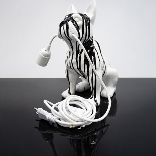Biała lampa w kształcie psa z czarnymi smugami, trzymająca w pyszczku białe przewody elektryczne z wtyczką, stojąca na czarnej, połyskliwej powierzchni.