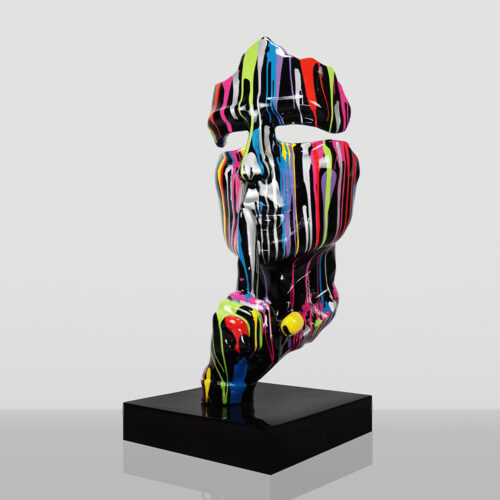 Rzeźba abstrakcyjnej twarzy z wyciętymi oczami, pomalowana czarną farbą z pionowymi kolorowymi smugami na biały, szarym tle, umieszczona na czarnej podstawce.