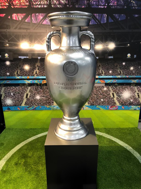 Symboliczne Trofea dla Euro 2024 3 Srebrny puchar UEFA European Football Championship ustawiony na czarnym podium na tle stadionu piłkarskiego z publicznością i oświetleniem.