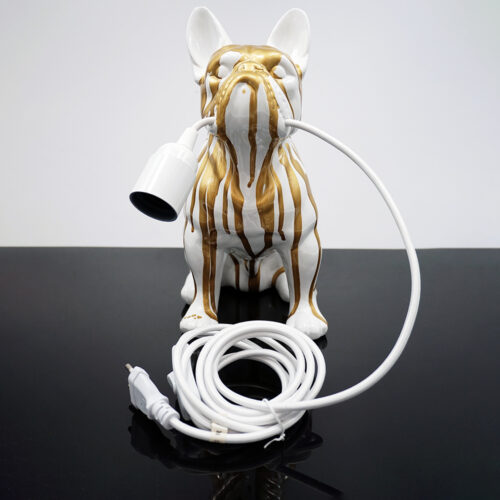 Biała ceramiczna figurka psa z pionowymi złotymi smugami, trzymająca w pysku białą oprawkę lampy z przewodem elektrycznym na czarnym połyskującym blacie