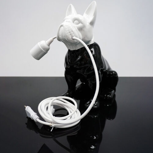 Lampka w kształcie psa buldoga w kolorze czarno-białym, trzymająca w pysku biały elastyczny kabel zakończony jasnym źródłem światła, stojąca na czarnej, błyszczącej powierzchni.