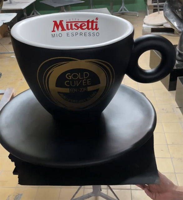 Figury Reklamowe Filżanek Musetti 1 Duża czarna filiżanka kawy z napisem Musetti Mio Espresso na białym wnętrzu oraz złotym napisem Gold Cuvée 1994-2014 na zewnątrz, ustawiona na czarnym spodku, na tle pomieszczenia z jasną podłogą i stołami.