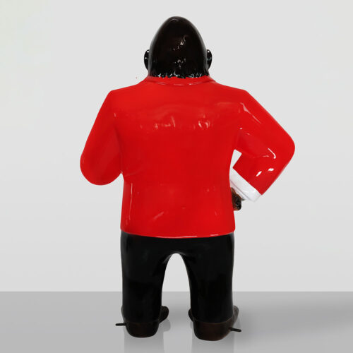 Figurka goryla widziana od tyłu, ubranego w czerwony żakiet, czarne spodnie i brązowe buty stojąca na szarej powierzchni przed jasnym tłem