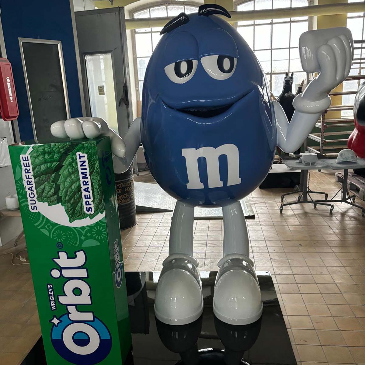 Niebieska figura z laminatu w kształcie M&M trzyma opakowanie gumy Orbit, stojąc w warsztatowym wnętrzu.