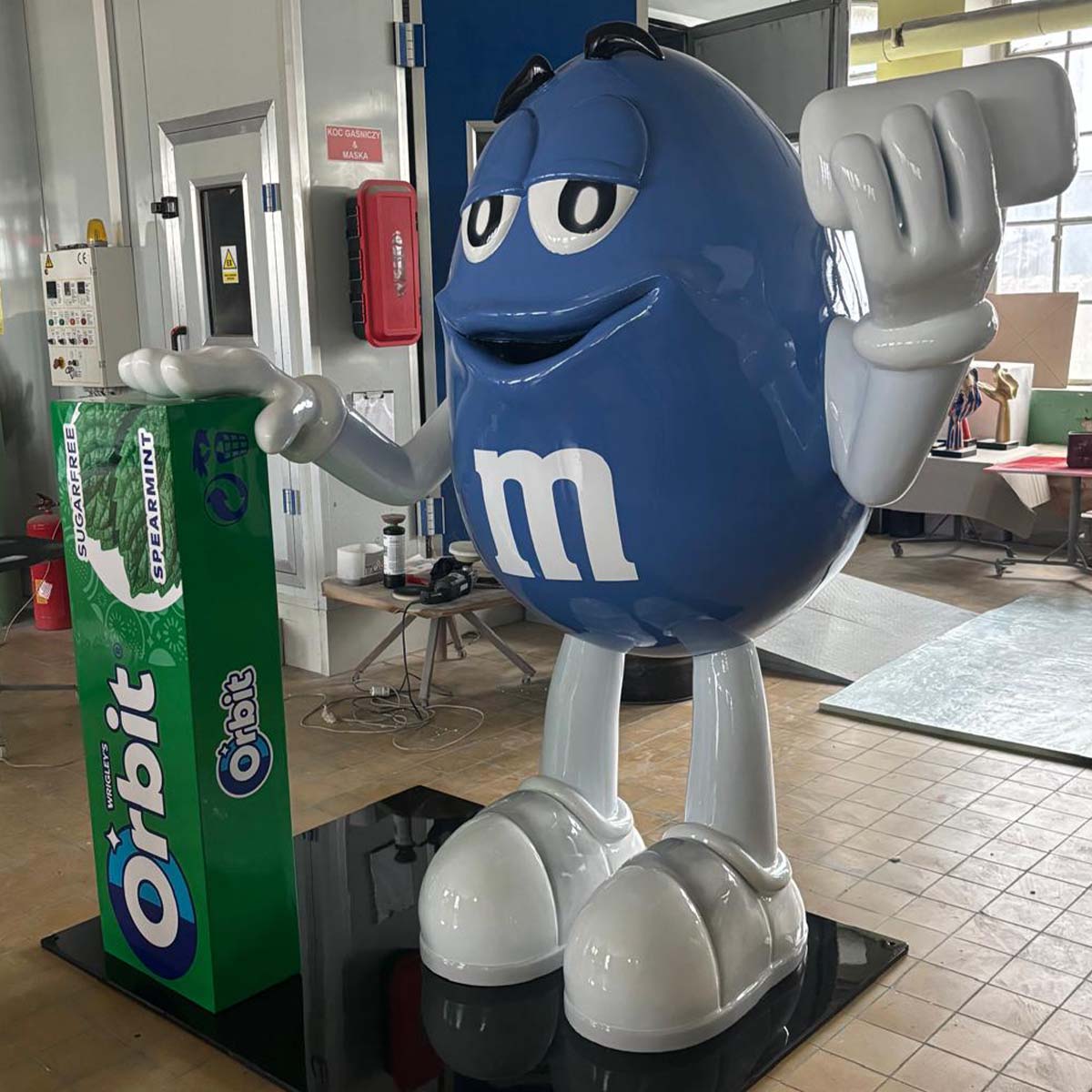 Niebieska figura z laminatu w kształcie maskotki M&M z białym „m”, stojąca obok zielonego stojaka Oribit w pracowni.