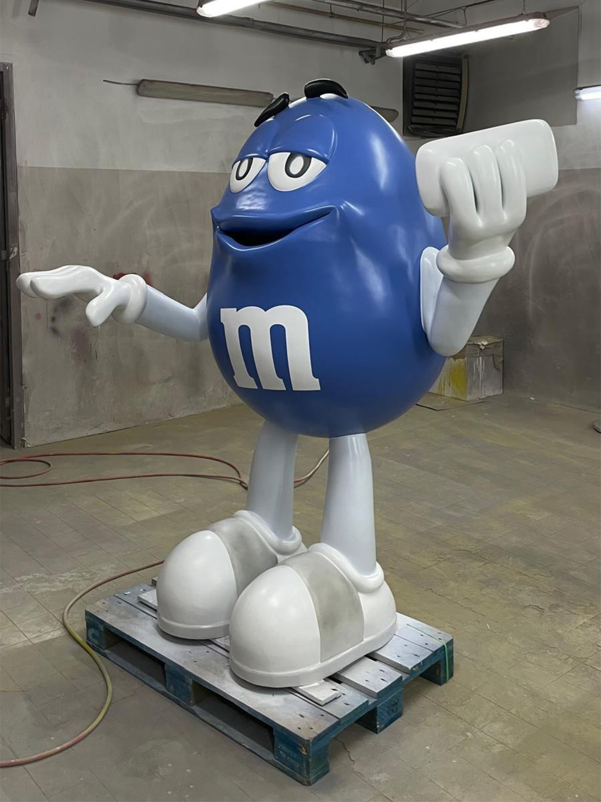 Niebieska figura z laminatu przedstawiająca postać M&M's z białymi rękami i butami, stojąca na palecie w warsztacie.