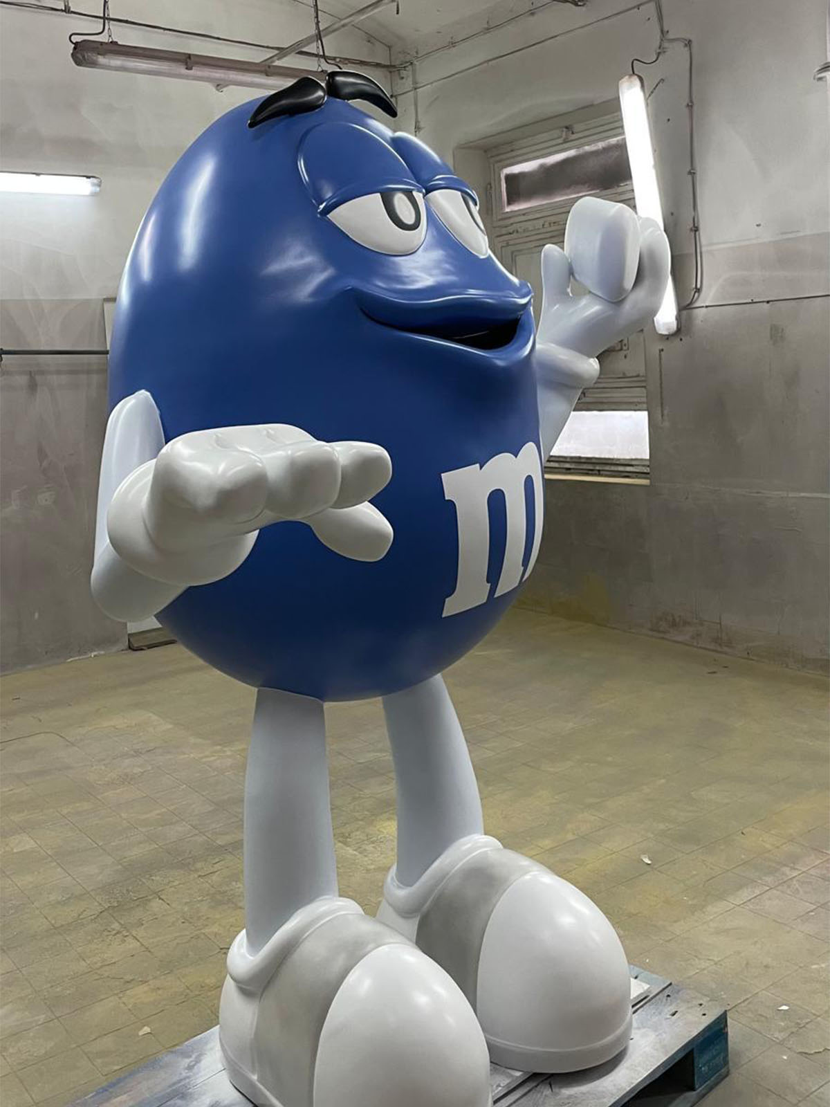 Wysoka figura reklamowa niebieskiej postaci M&M’s wykonana jako błyszcząca figura z laminatu stojąca w warsztatowym wnętrzu.