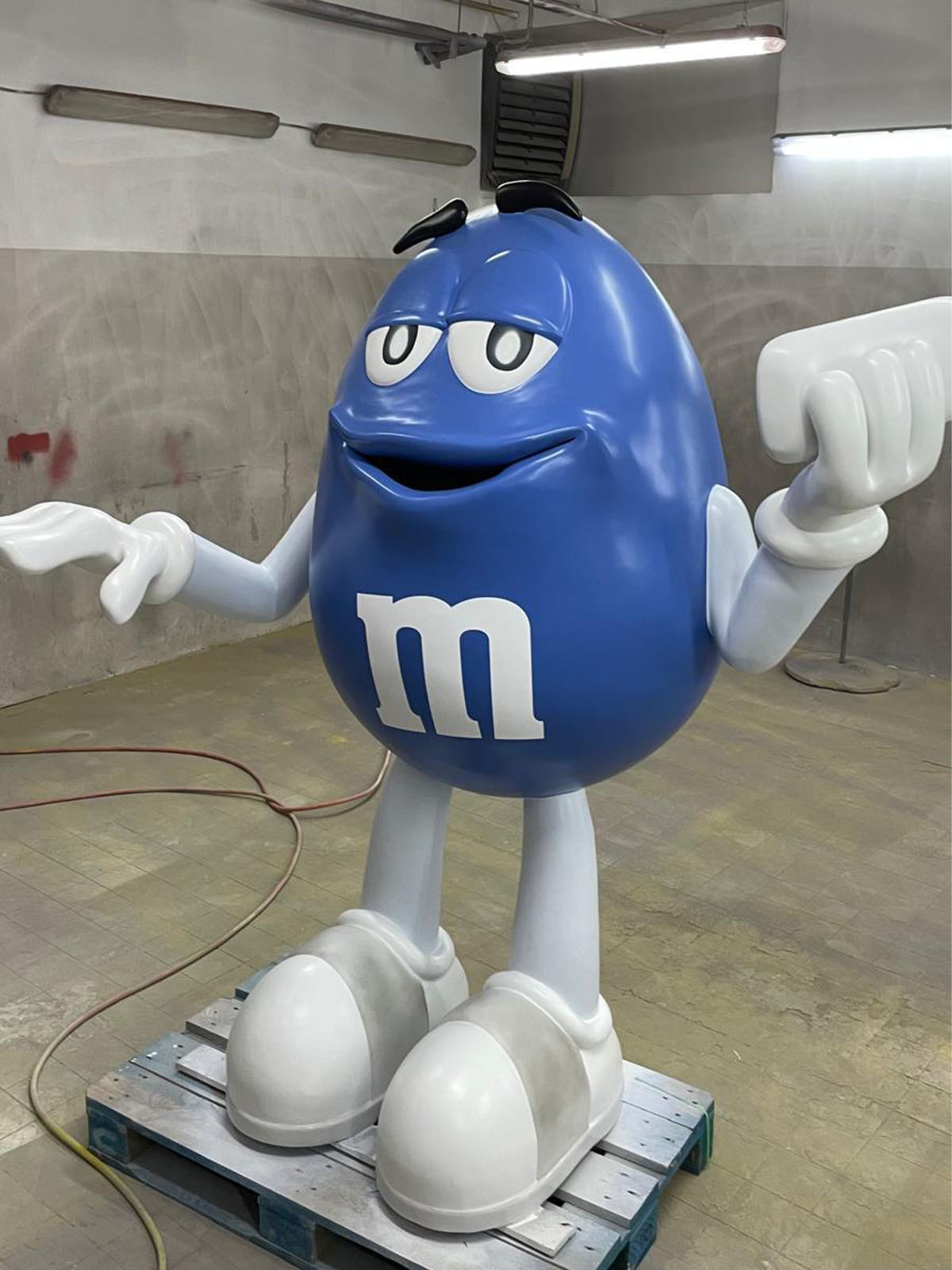 Duża niebieska figura z laminatu przedstawiająca postać M&M's z białym logo i uniesionymi ramionami, stojąca na palecie.