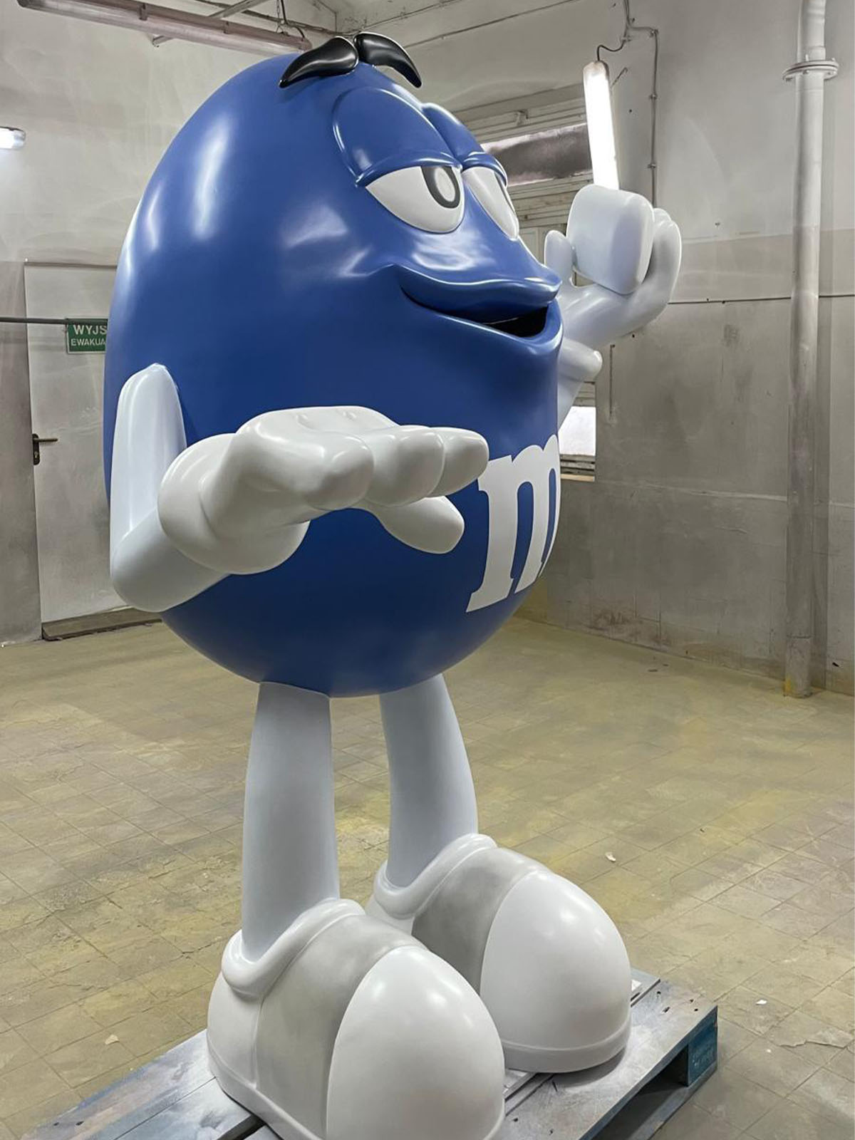 Niebieska figura z laminatu w kształcie maskotki M&M stojąca na dużych białych butach jako figura reklamowa.
