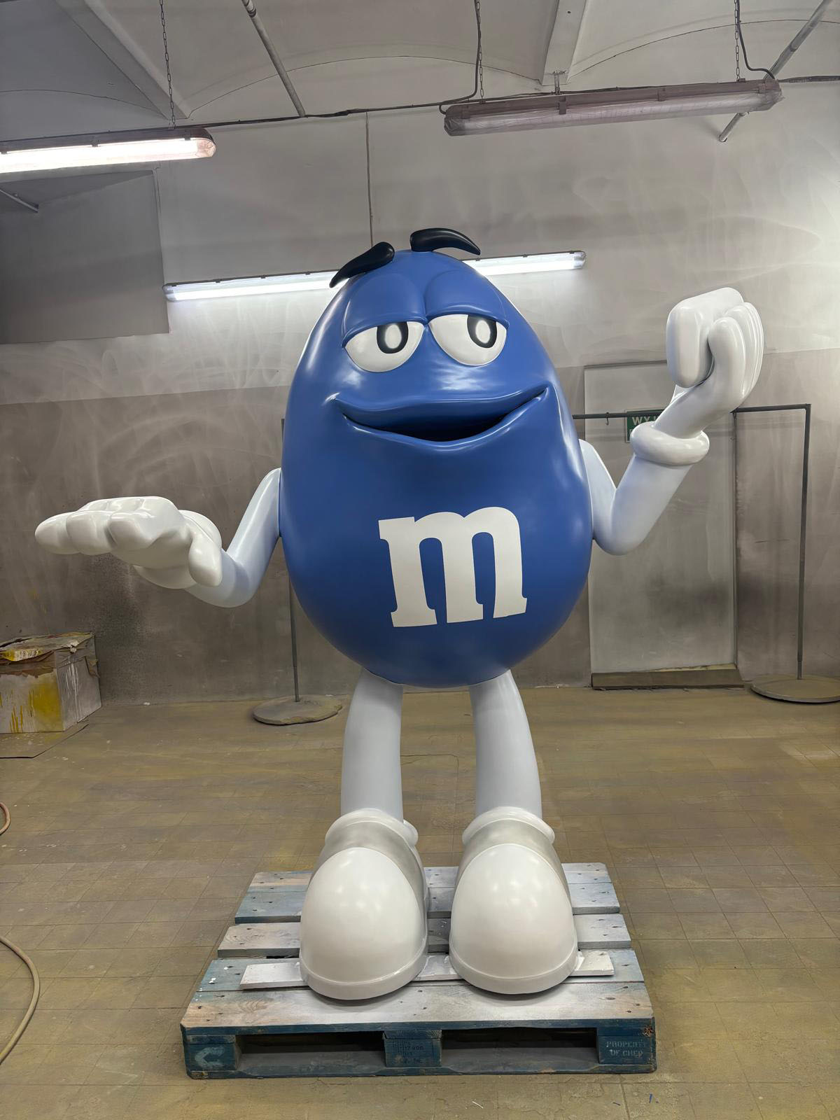 Niebieska figura z laminatu przedstawiająca maskotkę M&M’s w dużym rozmiarze stojąca na palecie w pracowni.