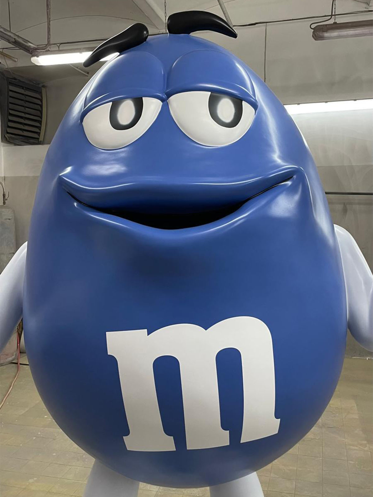 Duża niebieska figura z laminatu przedstawiająca maskotkę M&M z białym logo „m” i uśmiechniętą twarzą w magazynie.
