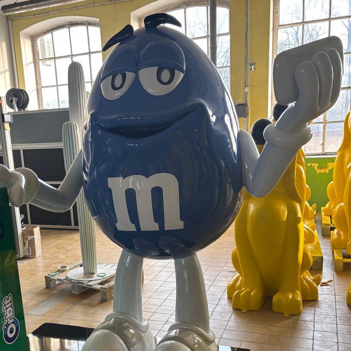 Niebieska figura z laminatu przedstawiająca postać M&M z unoszącą się ręką i wyrazistym logo na torsie.