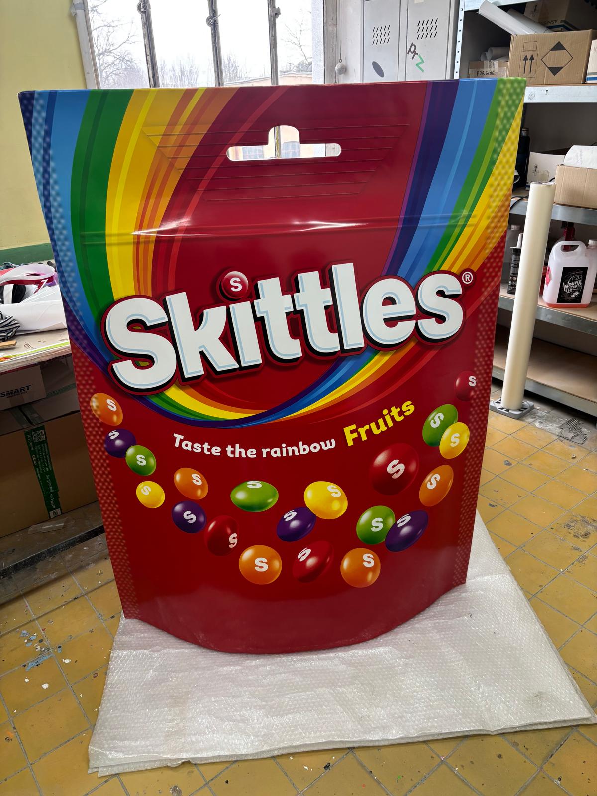 Duża figura reklamowa w kształcie opakowania Skittles z kolorową grafiką i rozsypanymi cukierkami, wykonana z laminatu.