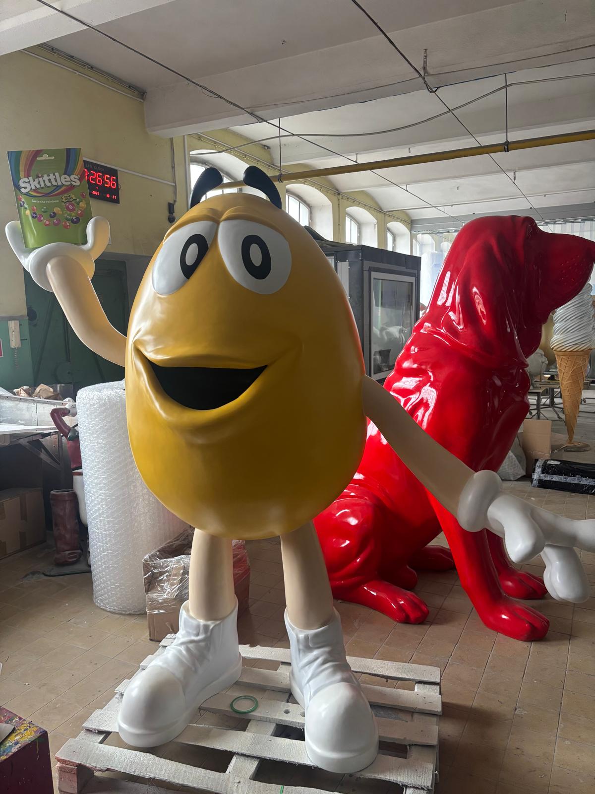 Żółta figura z laminatu w kształcie M&M trzyma opakowanie Skittles, stojąc w pracowni obok czerwonej rzeźby.