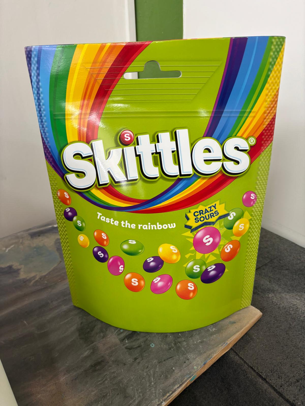 Duża zielona figura reklamowa w kształcie opakowania Skittles z kolorową grafiką i unoszącymi się drażetkami.