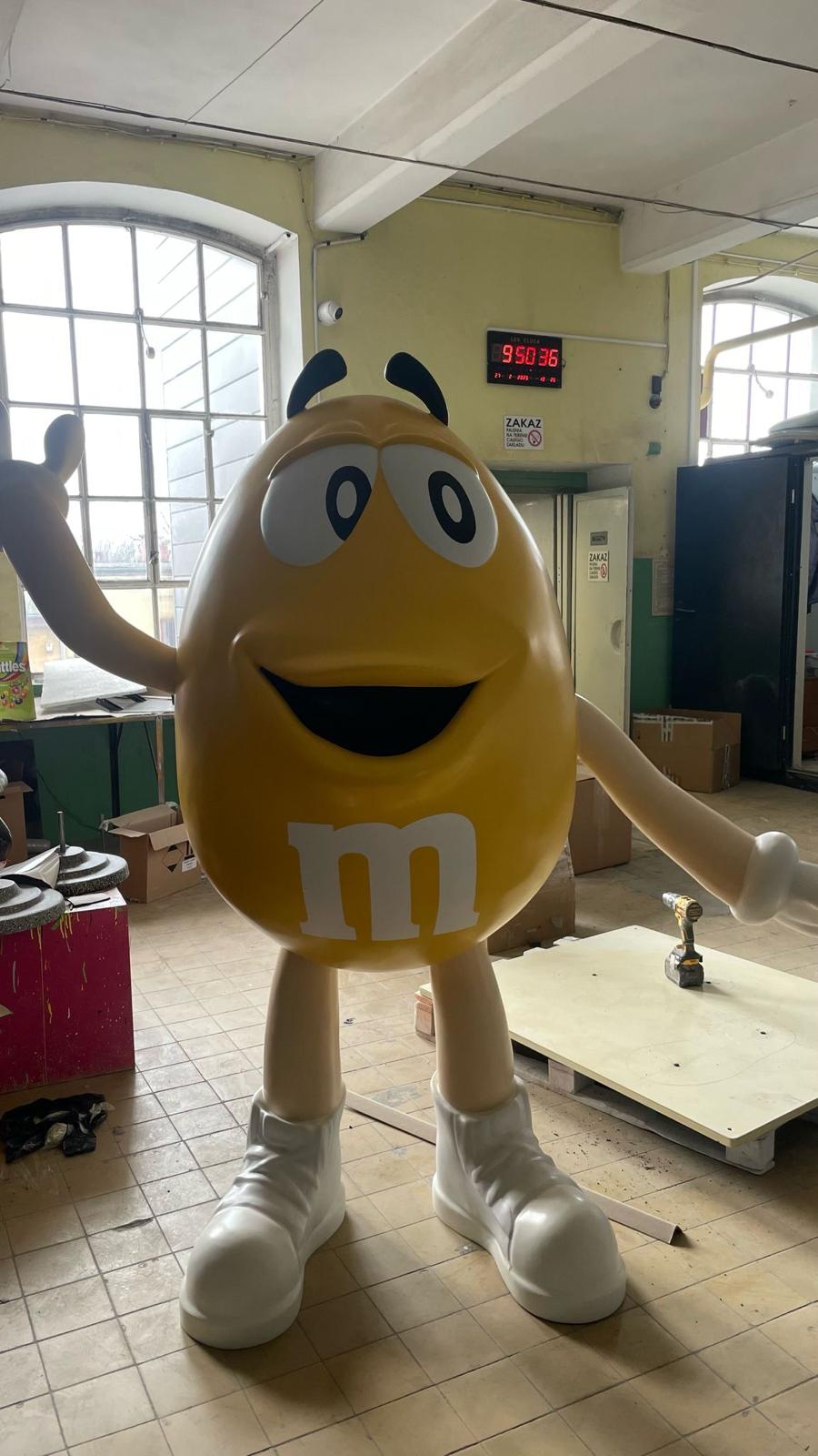 Duża żółta figura z laminatu przedstawiająca maskotkę M&M's z uśmiechem i uniesioną dłonią, stojąca w pracowni.