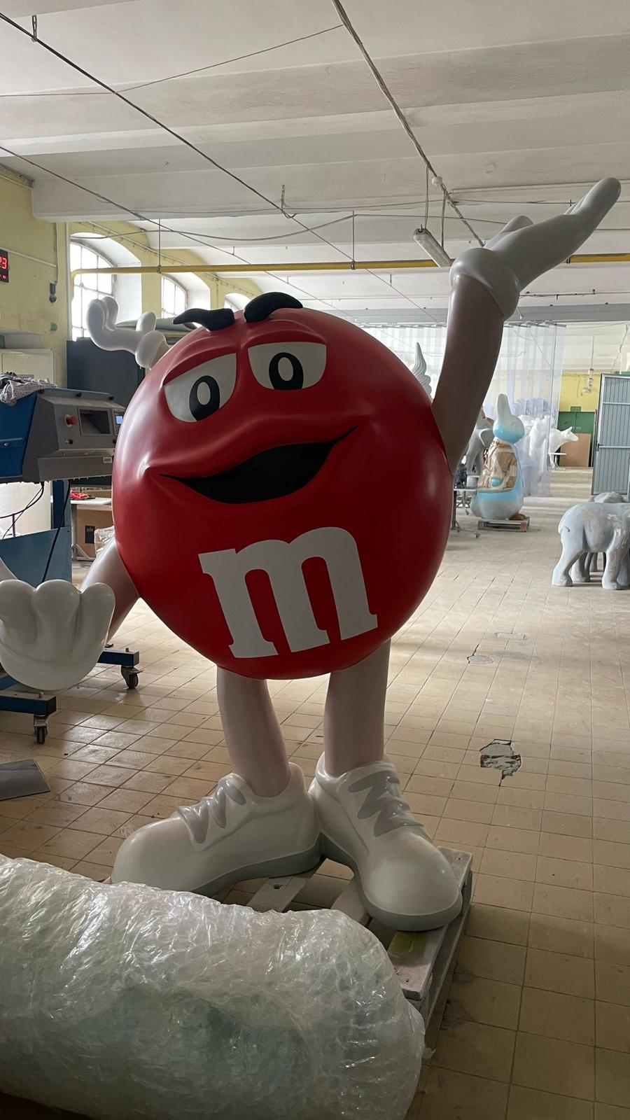 Duża czerwona figura z laminatu w kształcie M&M's z uniesioną ręką jako figura reklamowa ustawiona w pracowni.