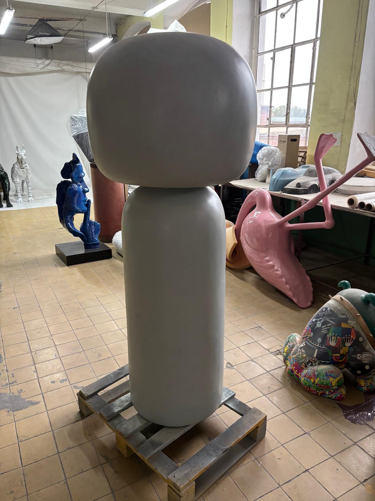 Wysoka prosta figura z laminatu o zaokrąglonej głowie stojąca na palecie w pracowni rzeźby artystycznej.